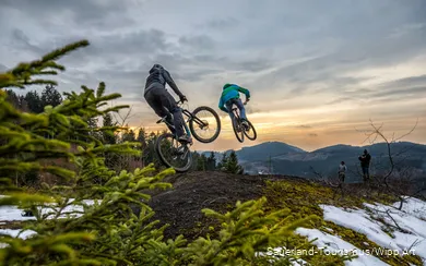 Zwei Mountainbiker springen über einen Hügel im Wald.