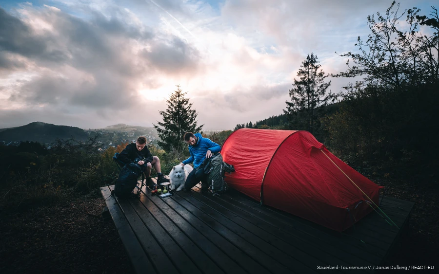 Zwei Wanderer und ein Hund bereiten das Lager für die Nacht auf einer Trekkingplattform vor.