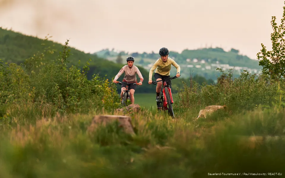 2 Mountainbikerinnen kommen einen Berg hoch, im Hintergrund sind Berge verschwommen zu sehen.