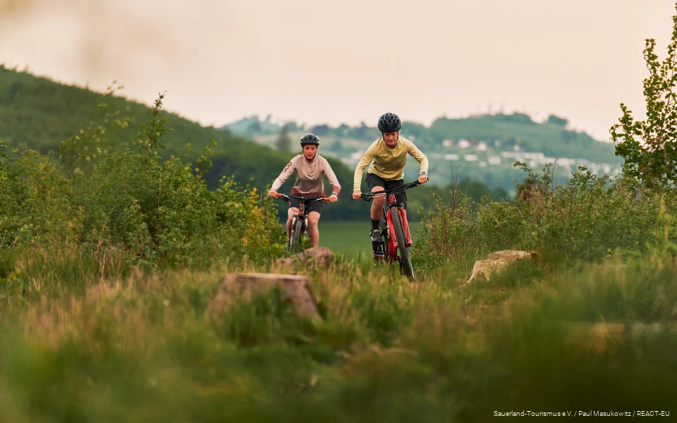 2 Mountainbikerinnen fahren über eine Wiese (2022) 2 Mountainbikerinnen kommen einen Berg hoch, im Hintergrund sind Berge verschwommen zu sehen.