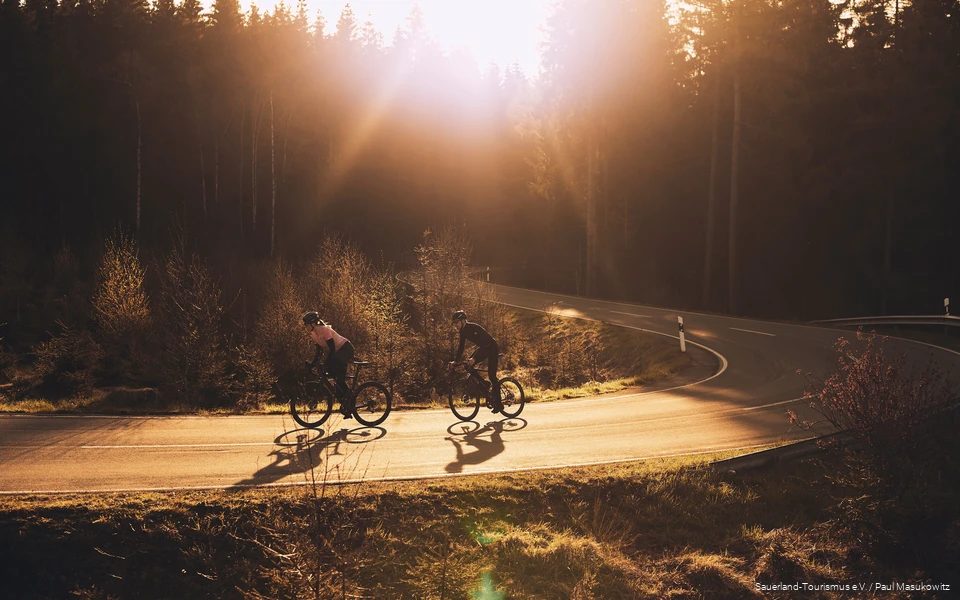2 Rennradfahrende fahren im Sonnenuntergang (weit) (2021) 2 Rennradfahrende fahren im Sonnenuntergang
