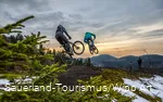 Zwei Mountainbiker springen über einen Hügel im Wald.